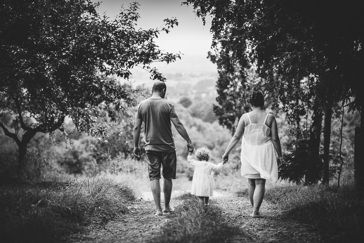 Czarno-biała fotografia rodzinna w lesie – rodzice prowadzący dwuletnią dziewczynkę leśną drogą, ujęcie od tyłu, naturalny spacer na sesji w Krakowie.